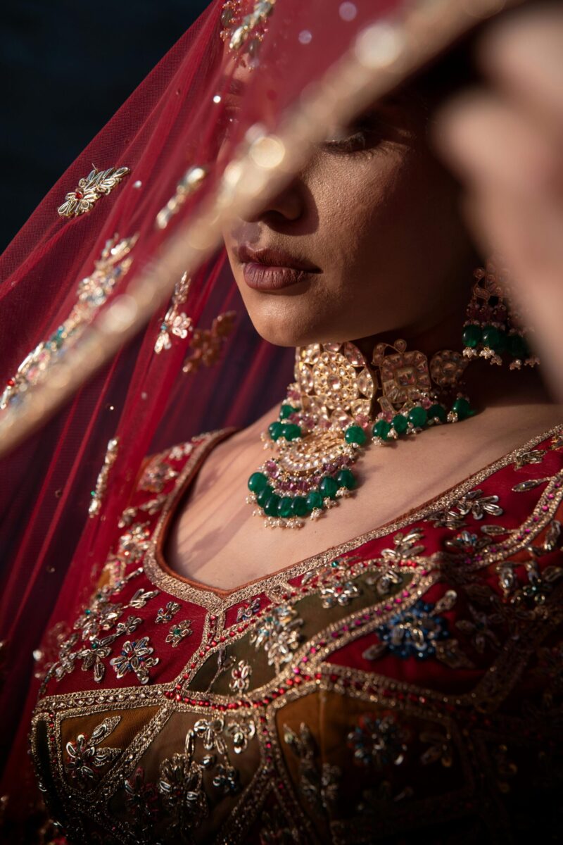 Asian bride in luxurious red and gold traditional dress with emerald jewelry and veil.
