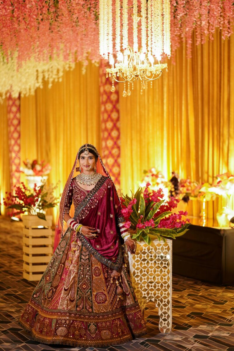 Stunning Indian bride posing indoors with floral decor and chandeliers, wearing traditional attire.
