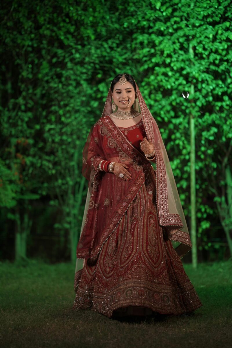 A joyful bride in a red lehenga poses outdoors during night-time wedding festivities.